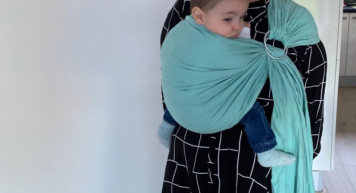 Kindje in een turquoise ringsling binnenshuis, het kijkt opzij
